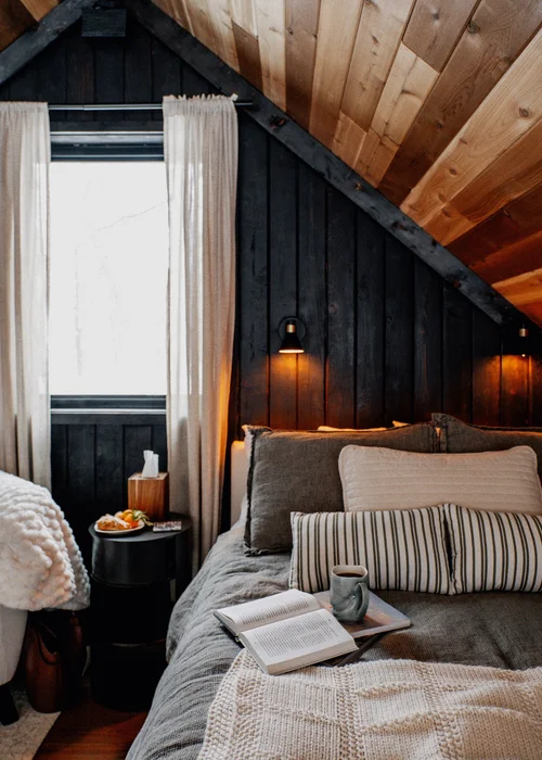 Cozy bedroom with a sloped wood-paneled ceiling, dark wood accent wall, and a large bed with multiple pillows. There is a bedside table with a plate of food, a book open on the bed, a mug, and a window with cream curtains.