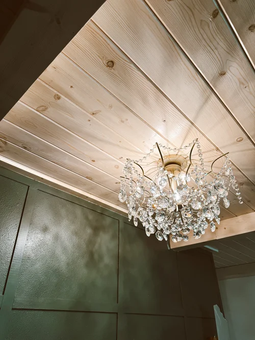 The image shows a crystal chandelier hanging from a wooden ceiling, with part of a textured green wall visible below.