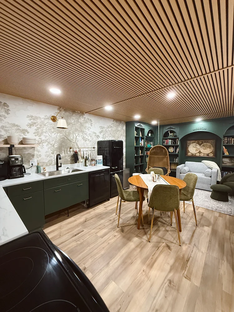 Interior of a cozy living space featuring a kitchenette with green cabinets, a round wooden dining table with six chairs, shelves filled with books, a white sofa, an armchair, and a wall with a mural of trees. The ceiling has wooden slats and recessed lighting.