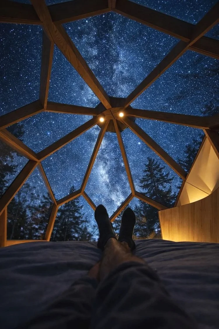 木造のジオデシックドーム屋根の建物の中でベッドに横たわり、天の川が輝く満天の星空を見上げている人物。